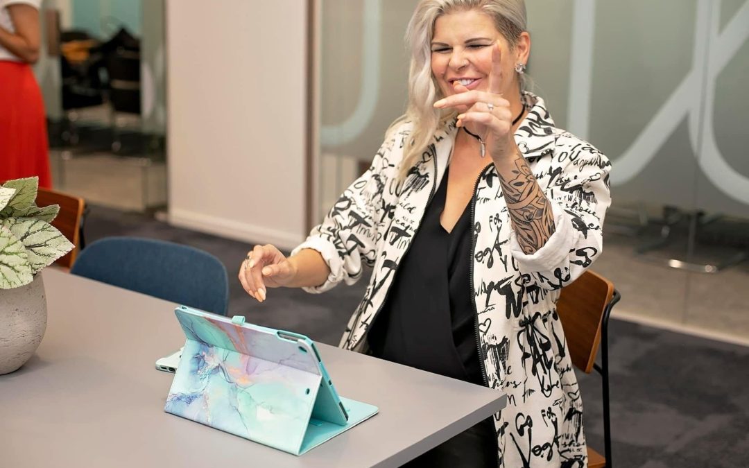 Charlotte Mather who runs the Hypnotherapy training at Axiom is pictured. She is a white female with long blonde hair, laughing as she points towards the camera. Charlotte is wearing a white Jacket with black and white writing and a black top.