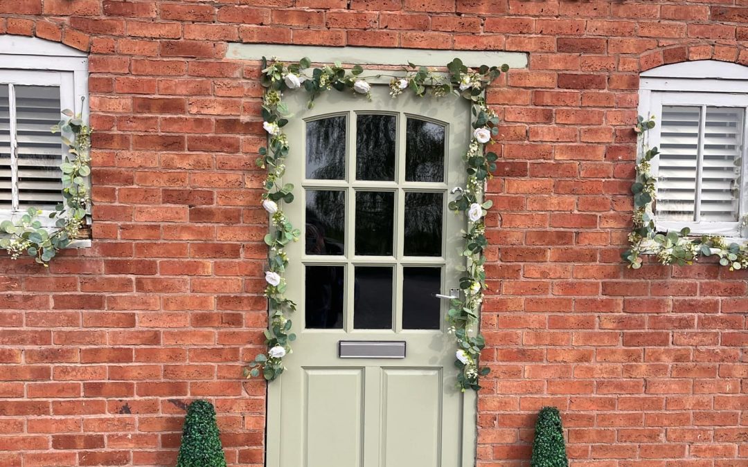 Front of the health barn hypnotherapy training centre, Sage green front door with a flower garland and a small evergreen tree either side. Two small windows on each side of the door with flower garlands