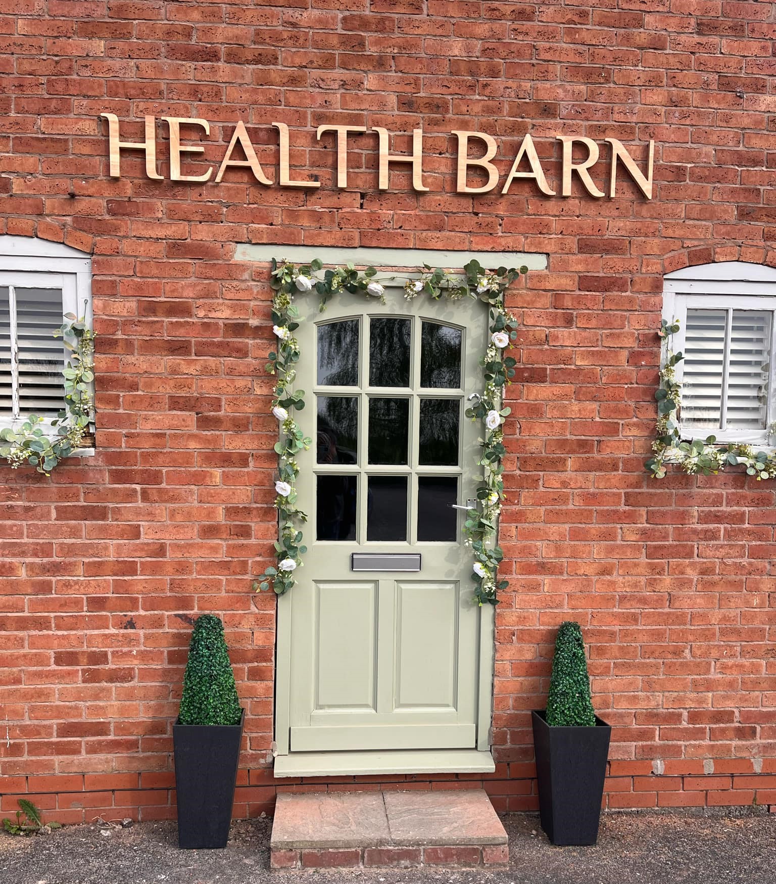 Front of the health barn hypnotherapy training centre, Sage green front door with a flower garland and a small evergreen tree either side. Two small windows on each side of the door with flower garlands